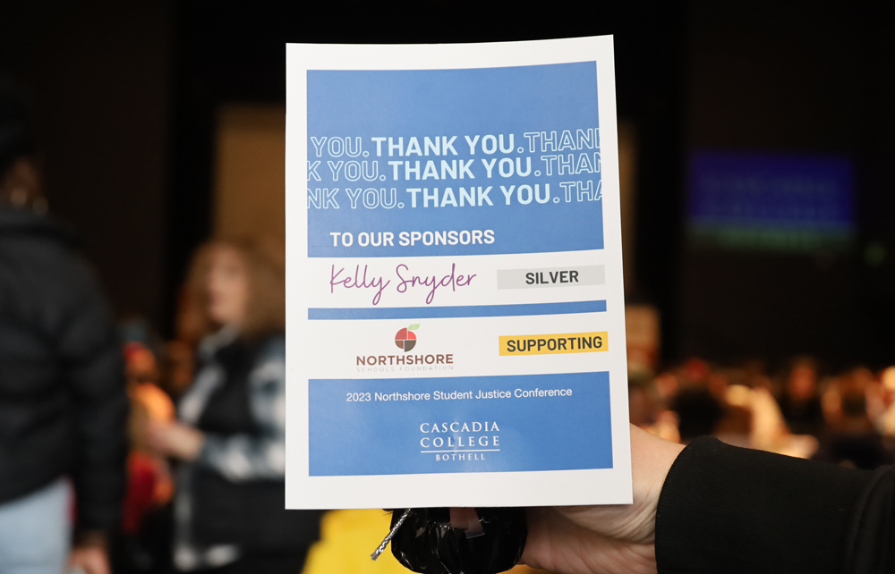 Sign in focus that reads, 'Thank you. Thank you. Thank you. To our sponsors Kelly Snyder, silver; Northshore Schools Foundation, supporting; 2023 Northshore Student Justice Conference' and Cascadia logo in front of an out of focus banquet crowd