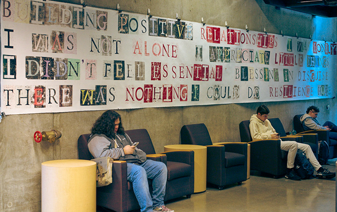 Students sitting by a wall with a large sign about positive relationships, looking at their phones.