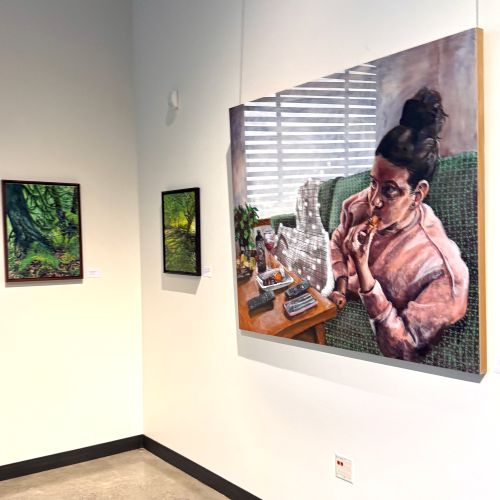 Corner of art gallery with two small tree landscapes and a large painting of a woman eating