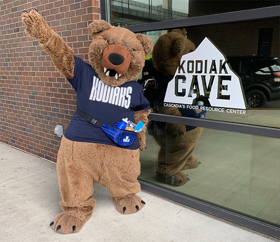 Cascadia College Kody mascot wearing Cascadia t-shirt and fanny pack waving in front of Kodiak Cave window